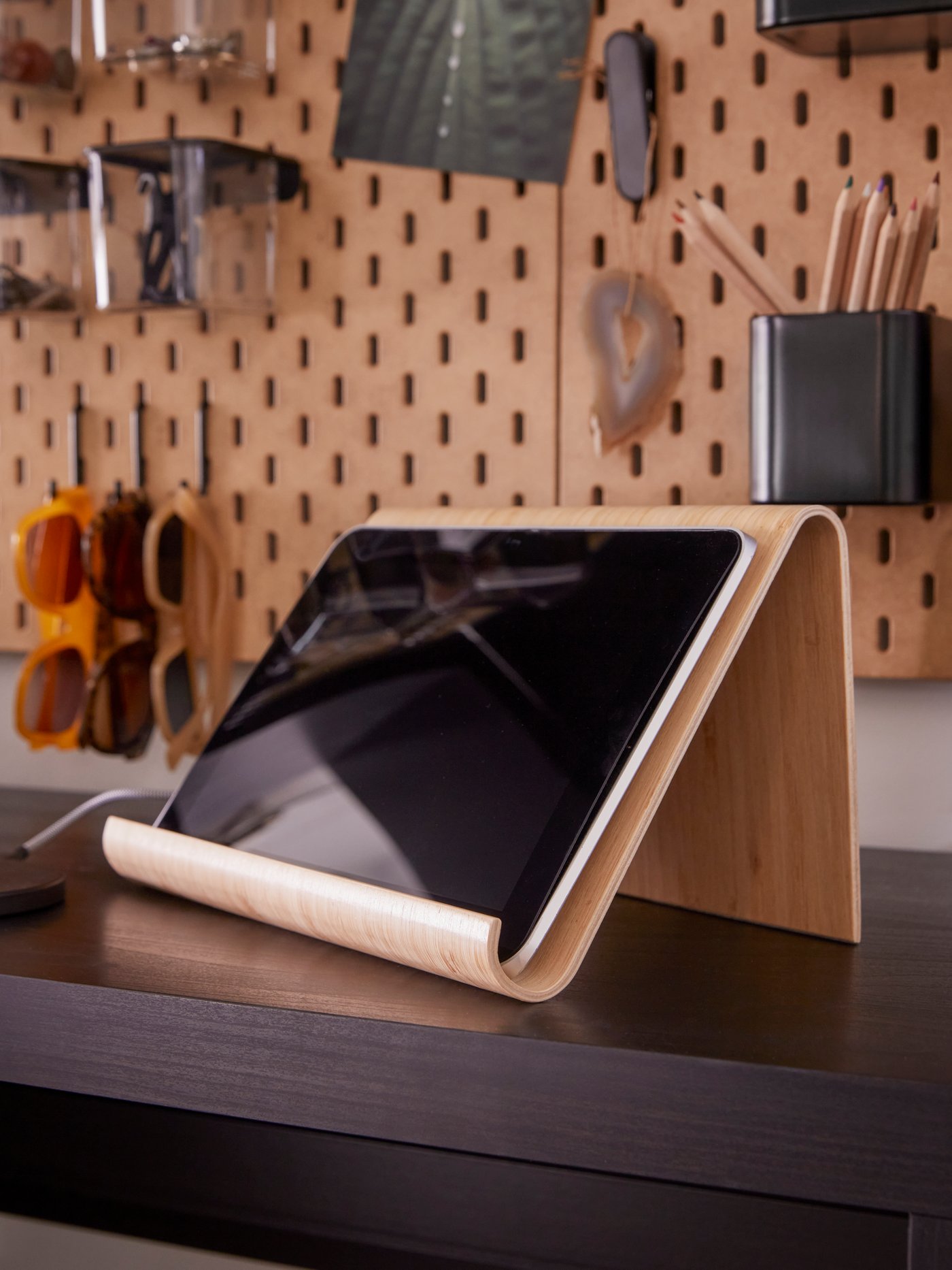 Držač za tablet VIVALLA od bambusovog furnira, sa tabletom na njemu, stoji na radnom stolu iznad kojeg visi perforirana tabla sa dodatnom opremom.