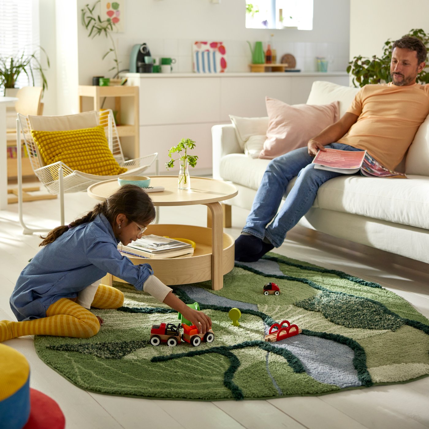 In a sunlit living room, a child sits on a GREJSIMOJS rug and plays with a train set while their parent sits on the sofa.