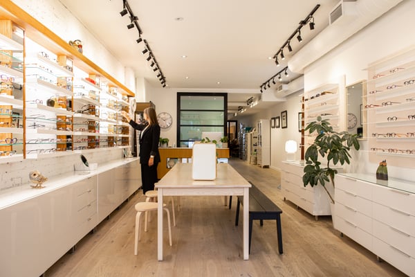 Bright optician's shop with long shelves filled with glasses, a central table and a person adjusting a product on the wall.
