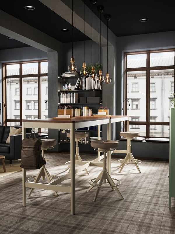 Two high IDÅSEN brown/beige tables placed close to a large window creating a standing meeting zone.