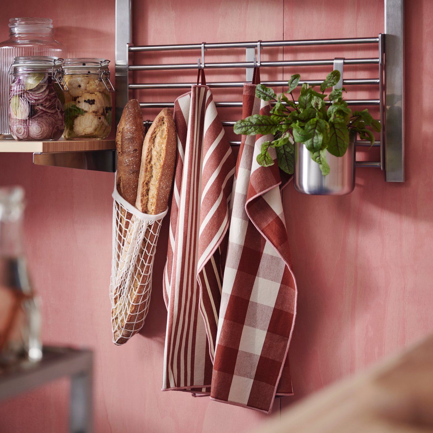 Two KÖSSEBÄR tea towels with mixed patterns, a mesh bag with bread, and a potted plant are hung on the rails of a wall shelf.