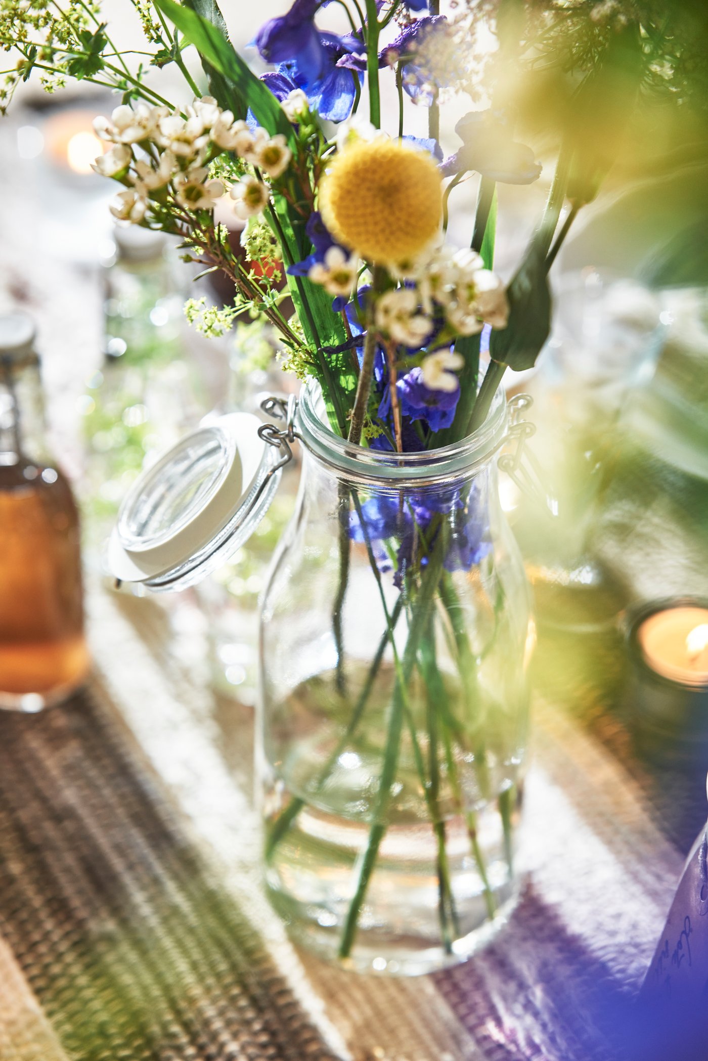 A glass bottle with colourful flowers on a table runner, with candlelight in the background.