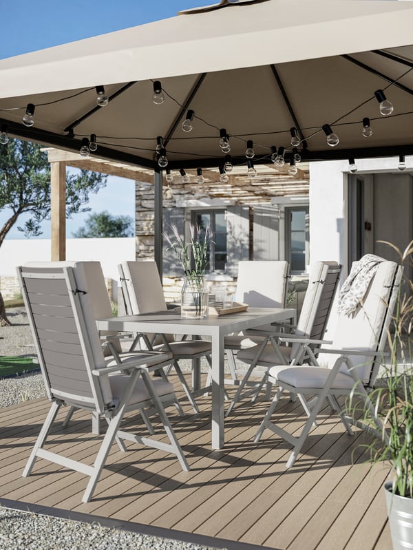 A dining table, outdoor chairs with cushions, wood-like decking, a grey-beige gazebo with hanging light chains.
