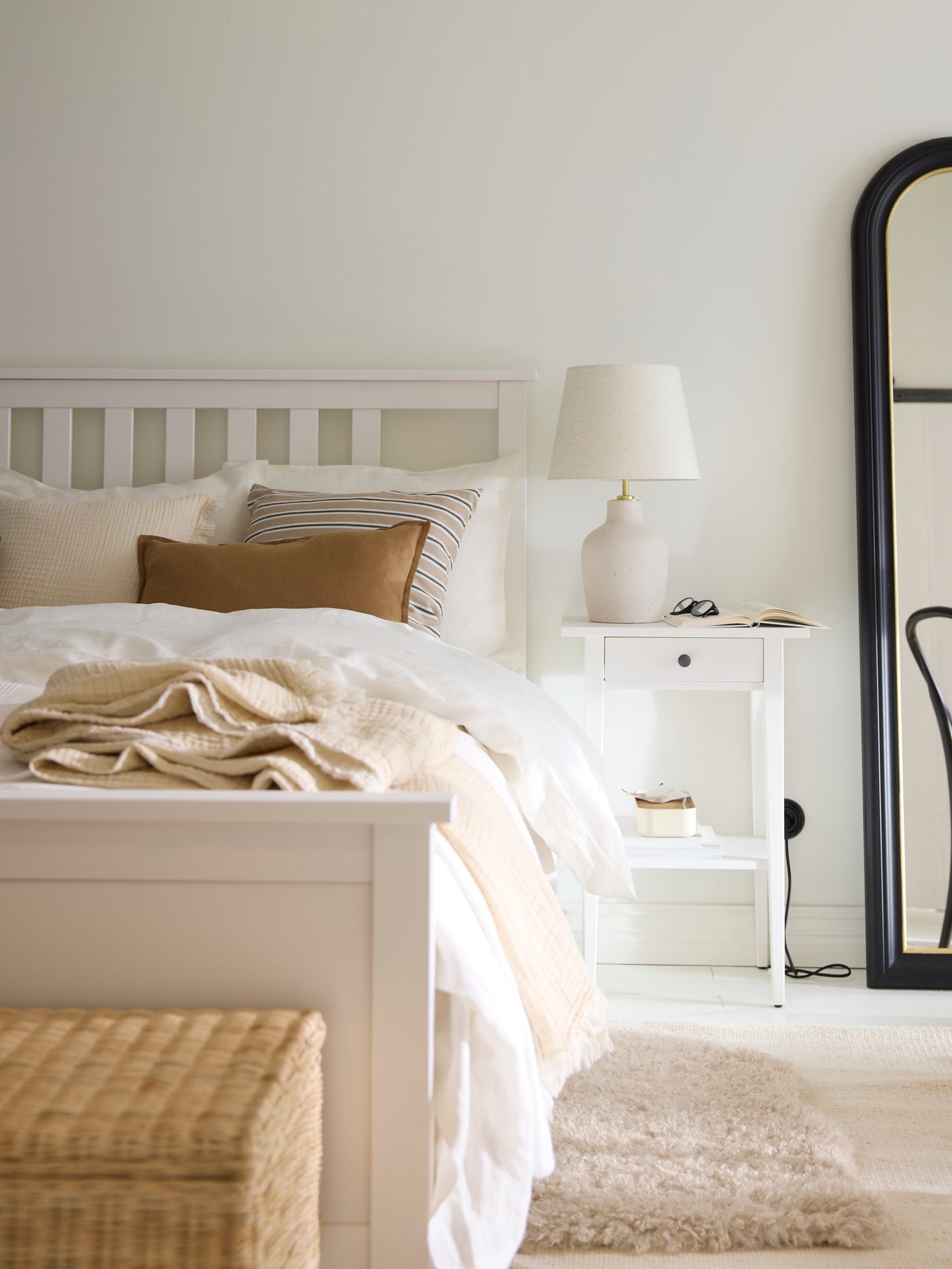 A white stain HEMNES bed covered in beige and white bed linen with a HEMNES bedside table and an ALMARÖD mirror beside it.