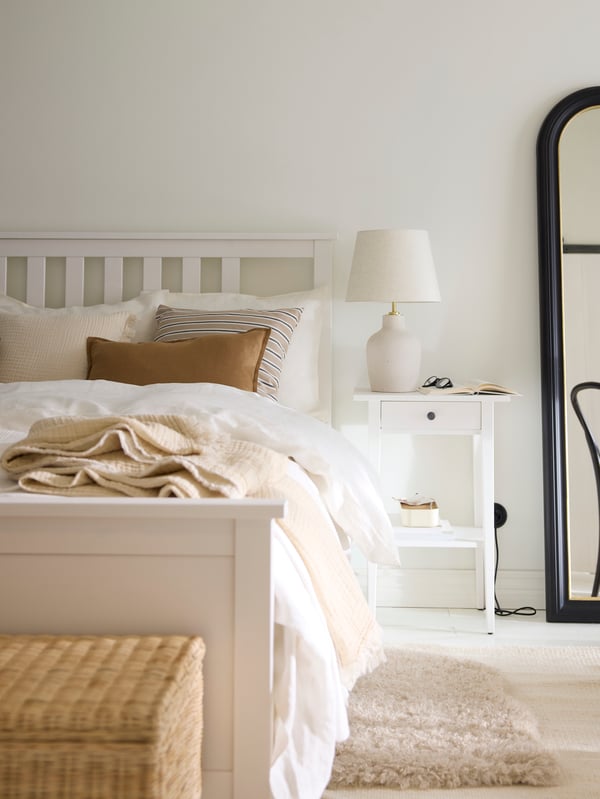A white stain HEMNES bed covered in beige and white bed linen with a HEMNES bedside table and an ALMARÖD mirror beside it.