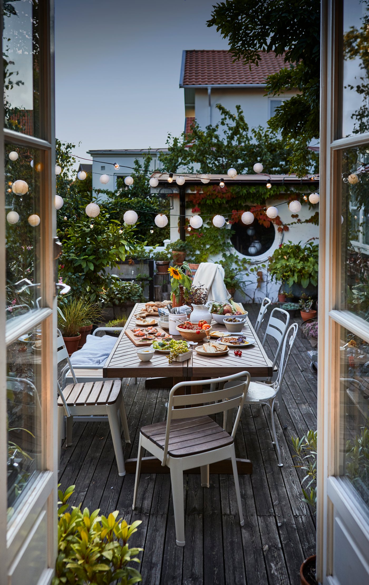 Mesa NORRMANSÖ para exterior en una terraza, rodeada de sillas y preparada para una comida al aire libre con vajilla y platillos servidos.