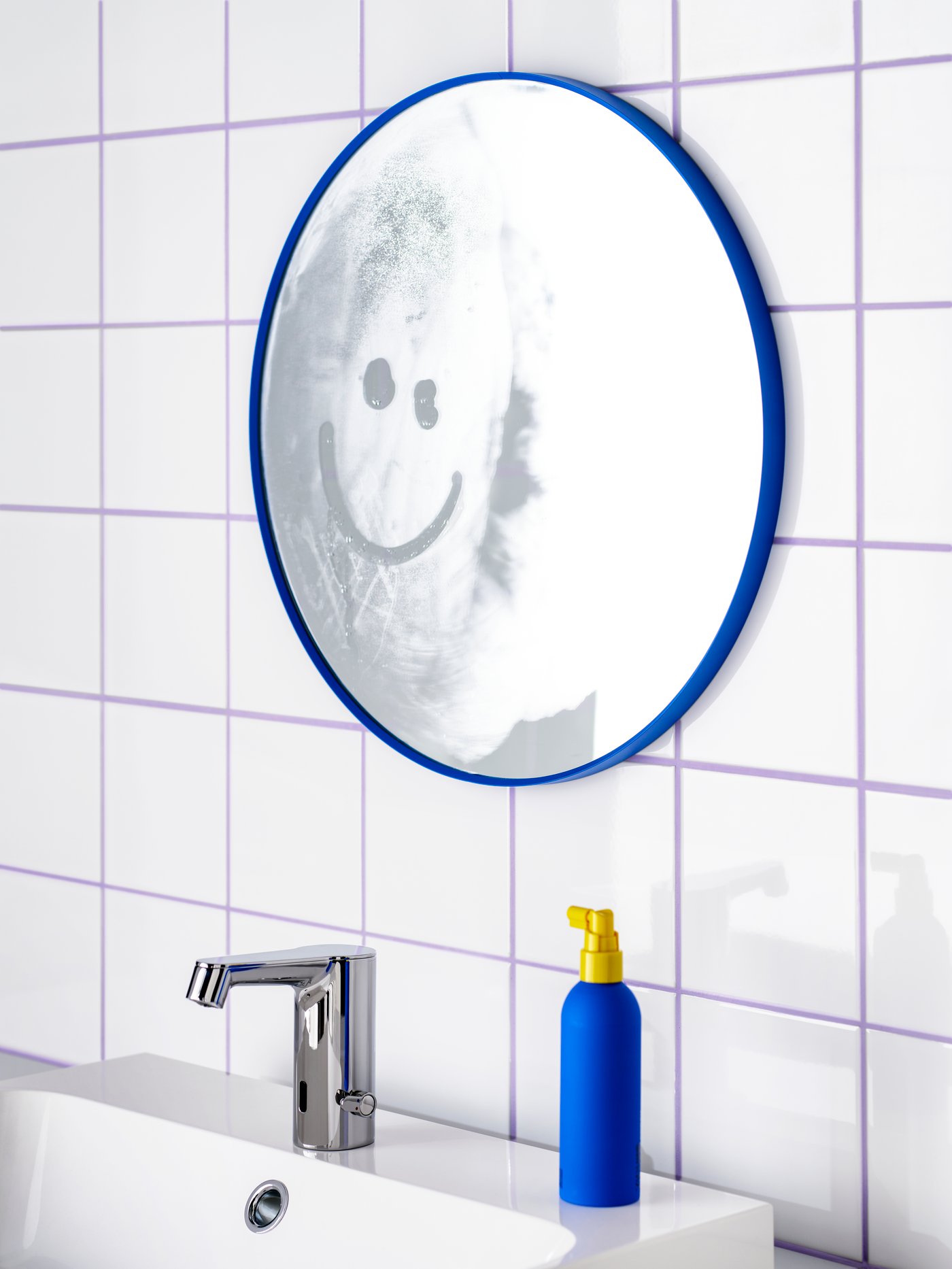 A blue LINDBYN mirror is mounted on the wall above a wash-basin. A smiley face has been drawn on the mirror’s steamy surface.