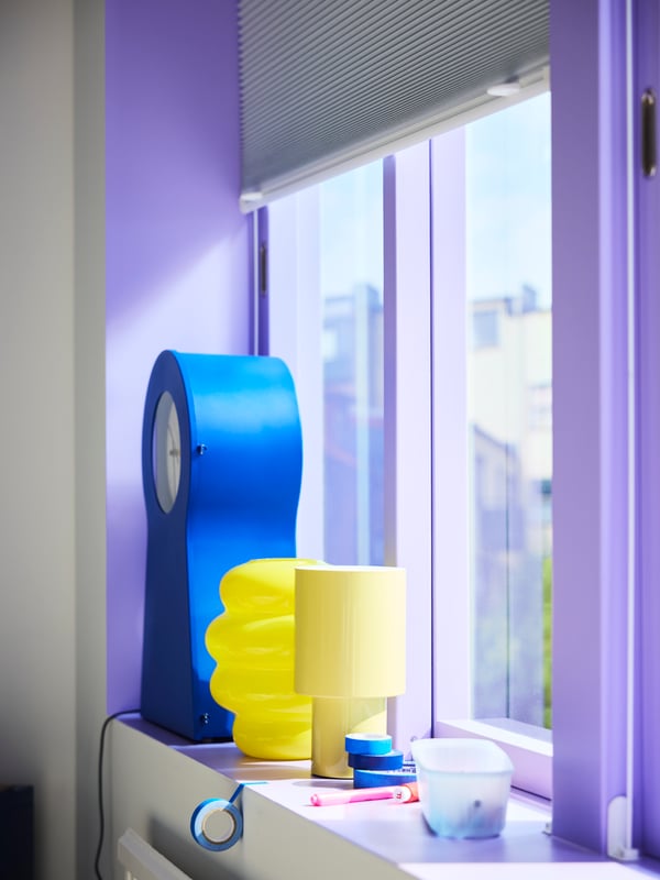 Close-up of a purple-painted window with bright yellow lamps and a blue IKEA PS 1995 clock standing on the windowsill.