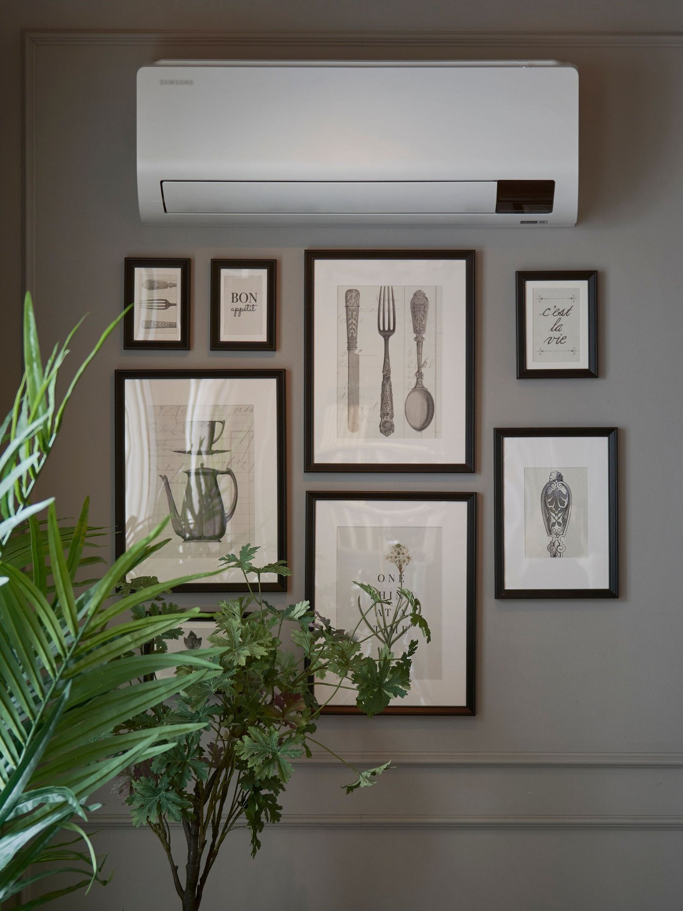 A wall with several framed black and white illustrations of kitchen utensils and plants, next to a green potted plant and under a white heat pump. The light comes from a window with light curtains on the left.