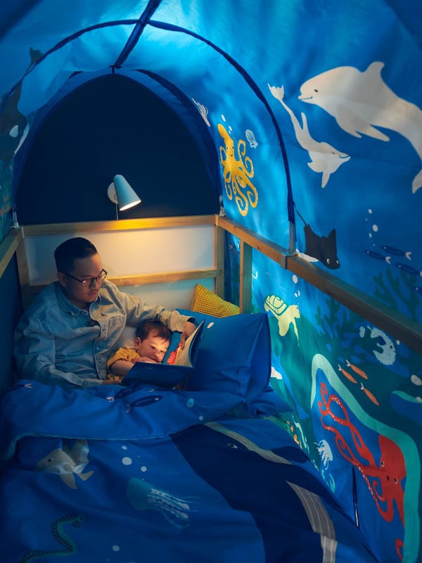 A man reads a bedtime story to a child in a KURA bed dressed in a BLÅVINGAD duvet set, covered by an ocean animal bed tent.