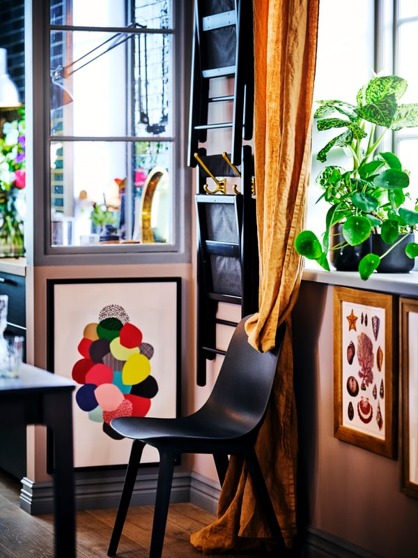 A black ODGER chair, surrounded by framed art on the walls, a folded TERJE chair and potted plants on the windowsill above.
