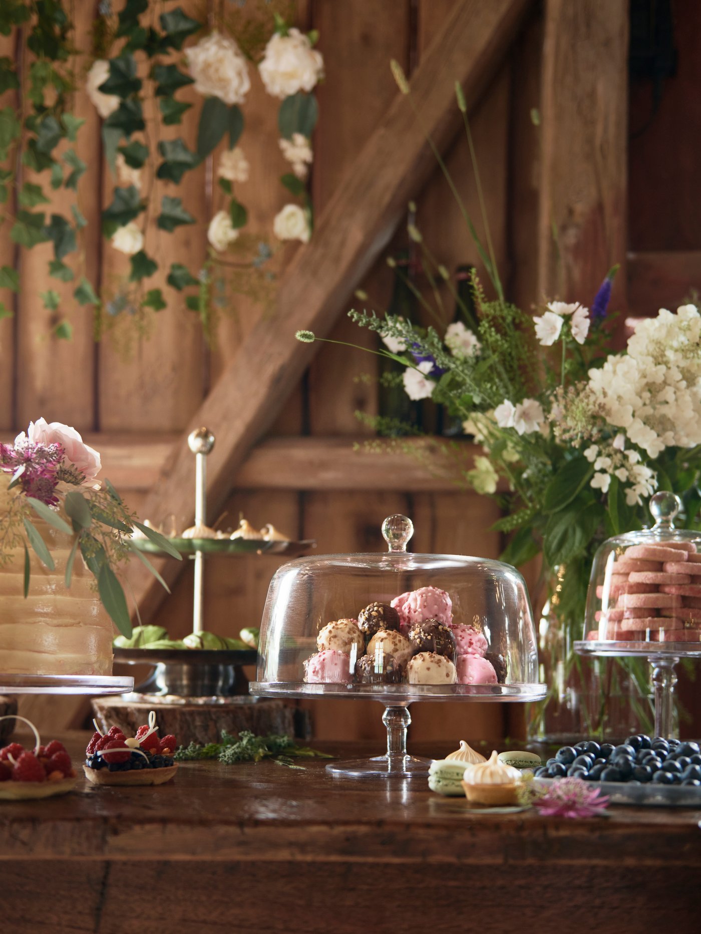 At a party, three INBJUDEN serving stands filled with sweets are set on a wooden table with flowers and other pastries.