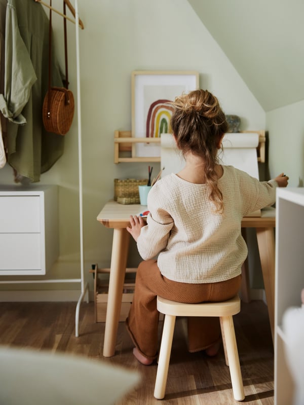 Fillette assise sur un tabouret pour enfants FLISAT en bois dessinant sur une table basse, à côté d’un portant blanc et d’une commode blanche.