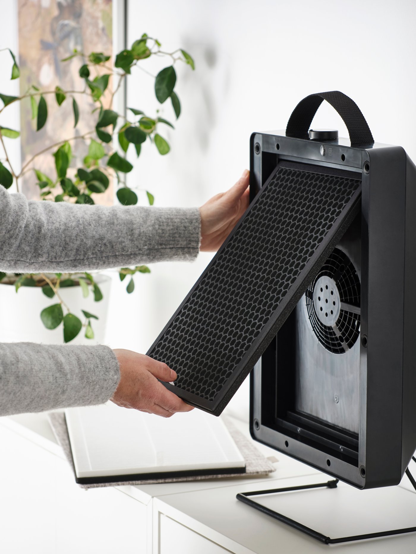 A person removing the gas filter from a black FÖRNUFTIG air purifier that is standing on a white chest of drawers.
