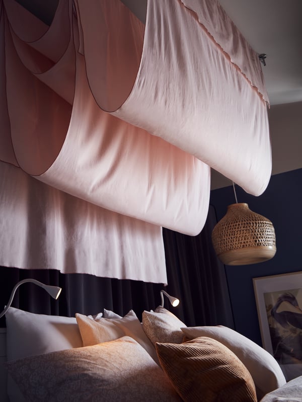 Lyserøde MOALISA gardiner bølger ned fra loftet ved siden af en loftlampe af bambus over en seng med beige/hvidt sengesæt.