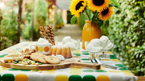 A cosy, summery set garden table with a green-yellow BRÖGGAN tablecloth. On the table are various treats, along with a yellow KOPPARBJÖRK vase containing yellow sunflowers.
