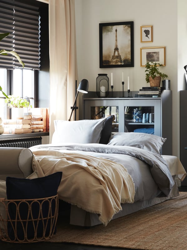 A light beige sofa-bed made as a bed with grey bedding. Behind is a grey glass-door cabinet and a jute rug on the floor.
