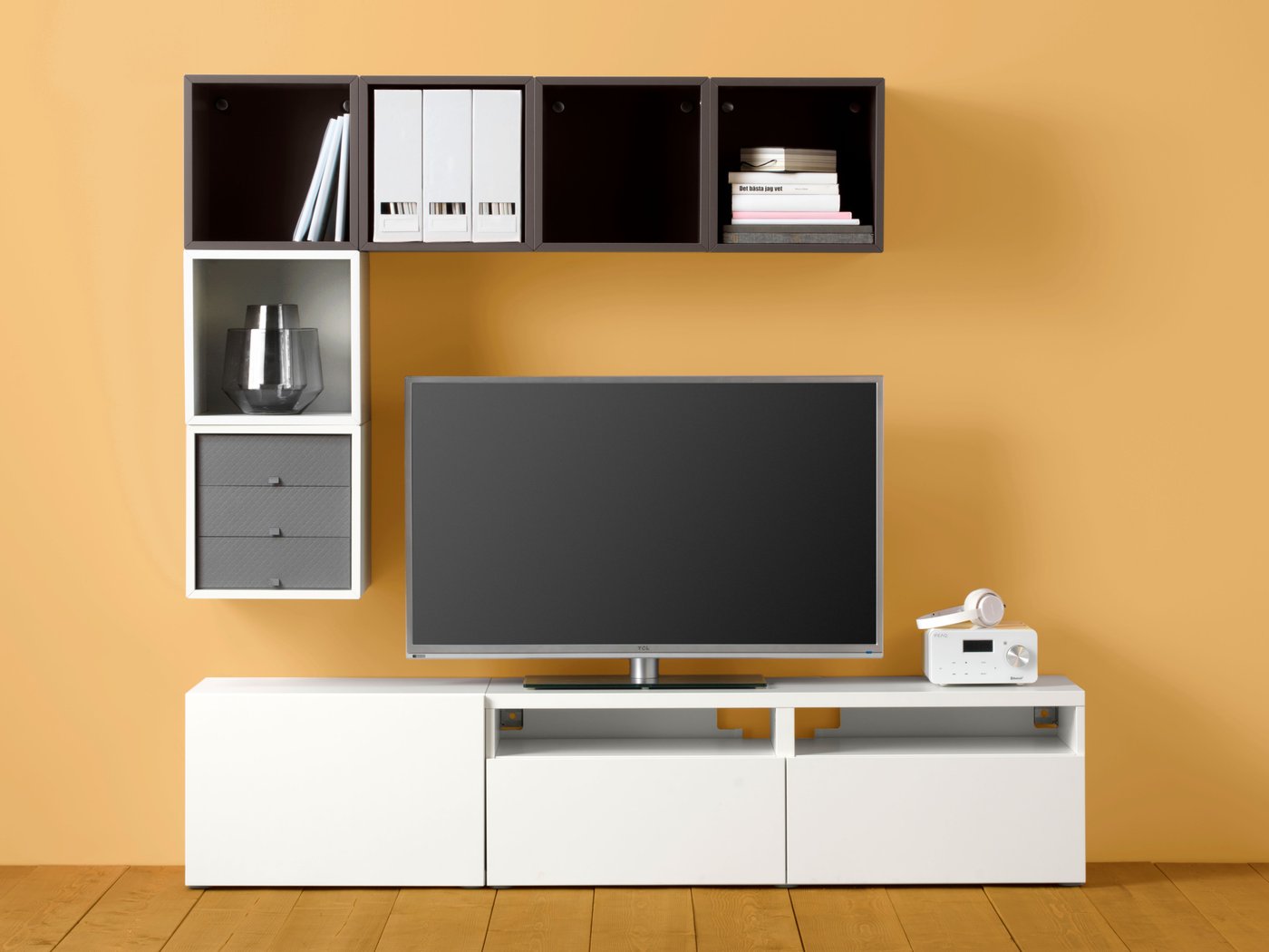 A TV framed top, side and bottom by a geometric but asymmetric combination of BESTÅ and EKET cabinets against a yellow wall.