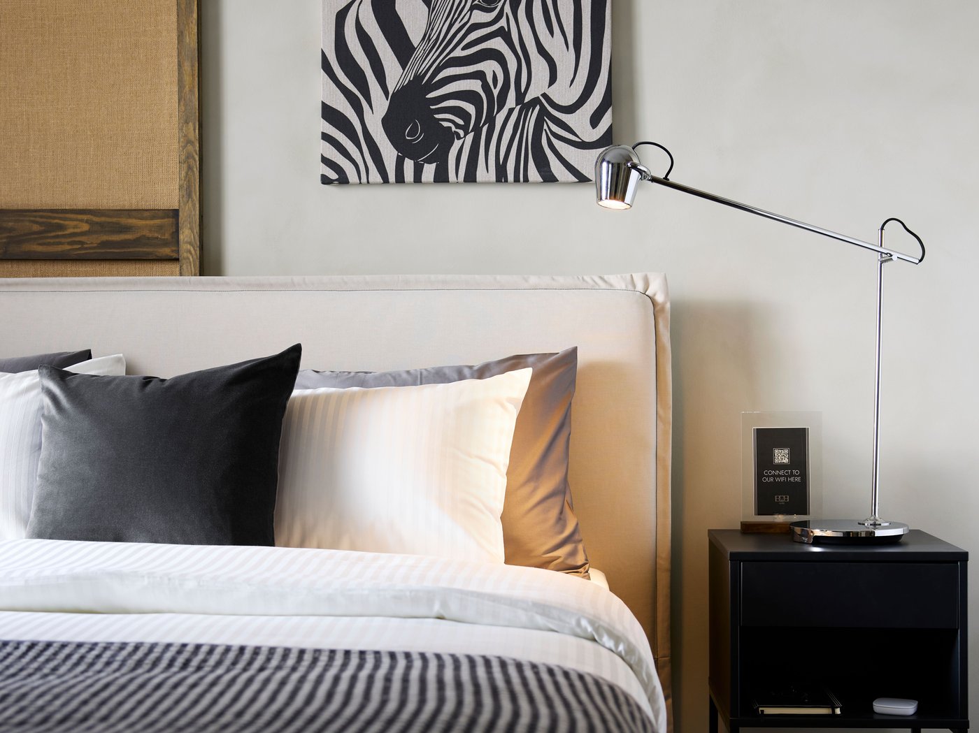 Ein beigefarbenes gepolstertes TÄRNKULLEN Bettgestell setzt in einem Hotelzimmer einen schönen Akzent. Das Bett ist frisch bezogen und steht neben einem Ablagetisch an einer Wand mit einem Kunstwerk mit Zebralook.