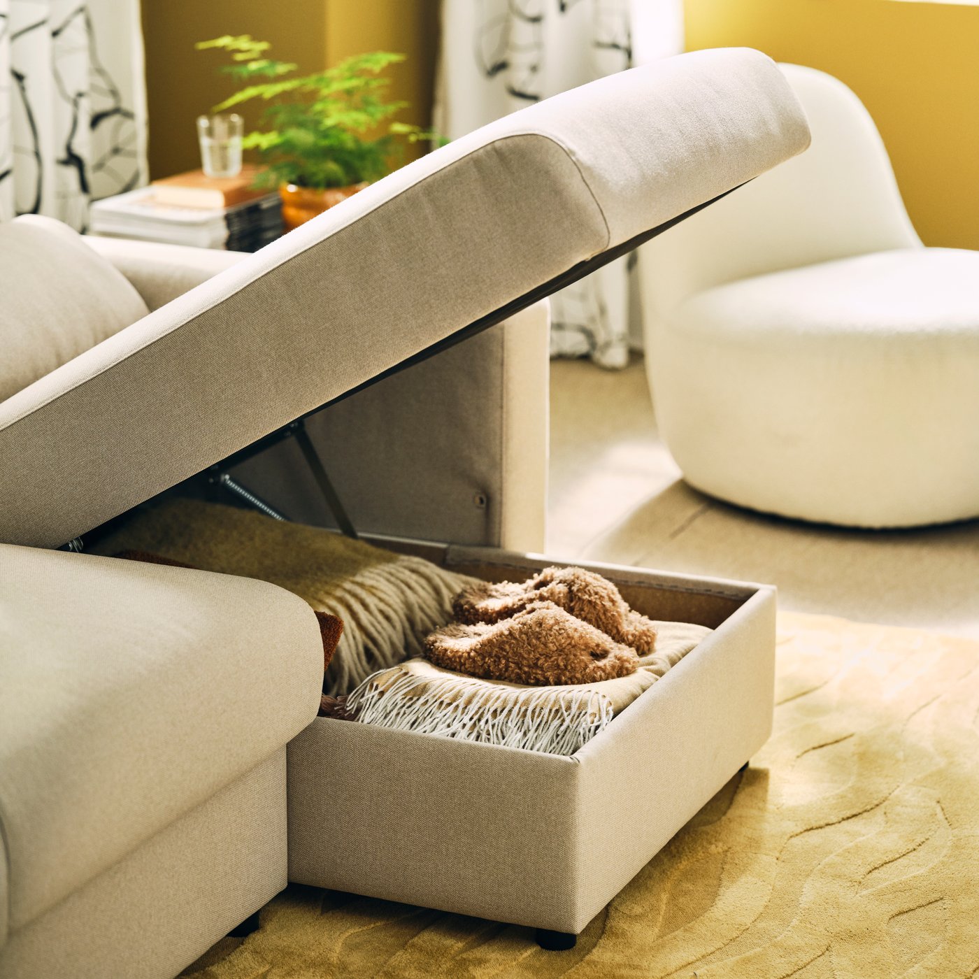A living room with a light-beige MALMKÖPING three-seat sofa with its chaise longue open, revealing stored textiles.