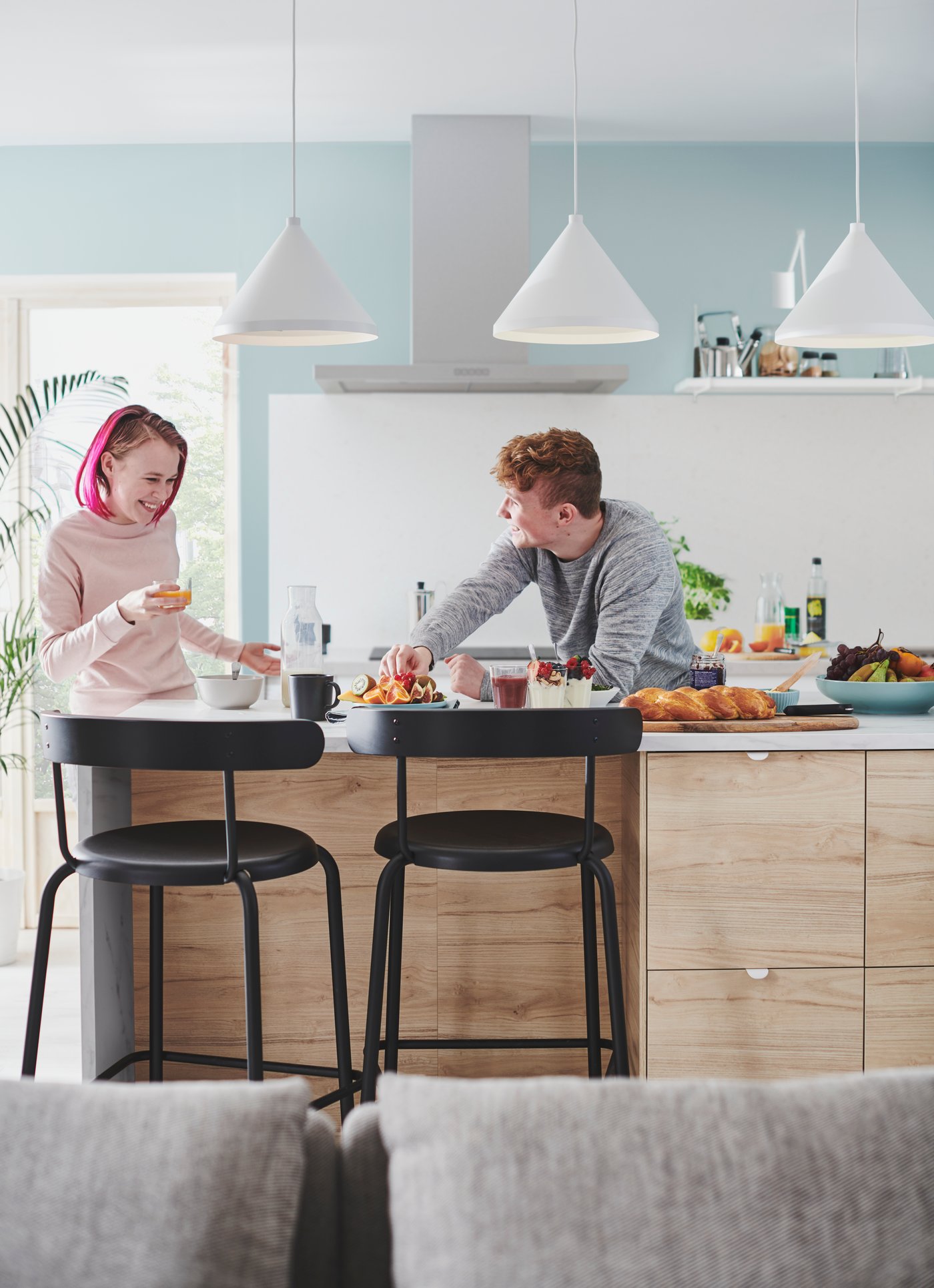 A man and a woman are standing behind a kitchen island, tasting the freshly cooked food together and laughing.