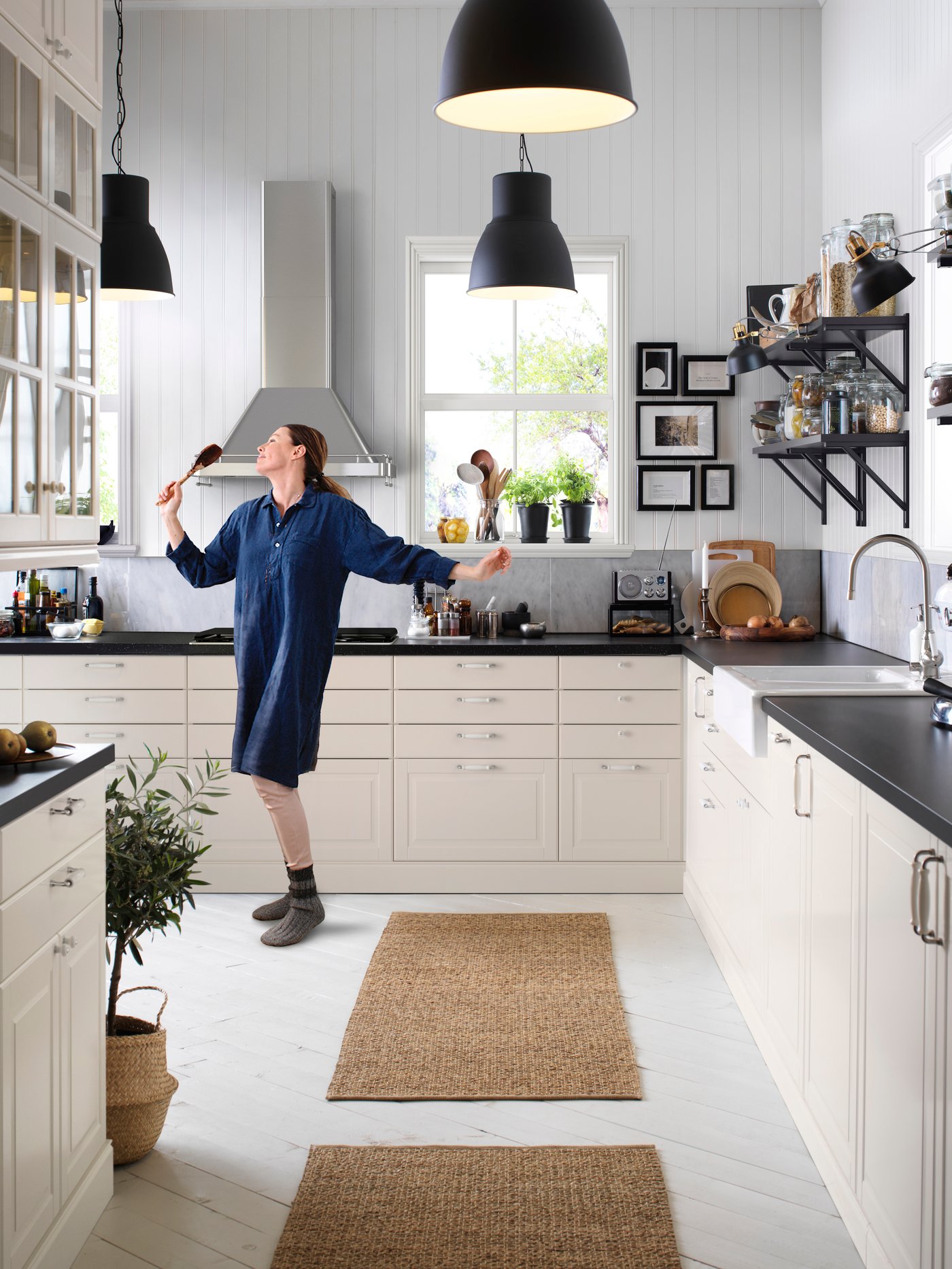 Modernt kök med vita skåp, svarta bänkar, två hänglampor, person med köksredskap, fönster och öppna hyllor.