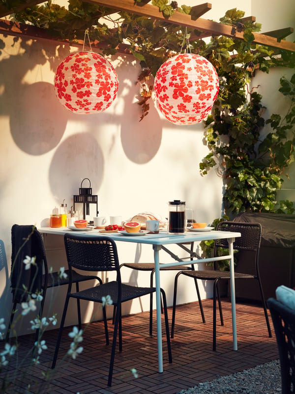 Hanging paper lanterns with a red floral pattern, a table set with breakfast and four black stackable chairs.