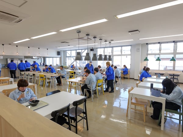 大きな窓と長テーブルが並ぶ休憩スペース。ブルーの制服を着た人たちがテーブルに座り、床は光沢のあるフロア仕上げ。