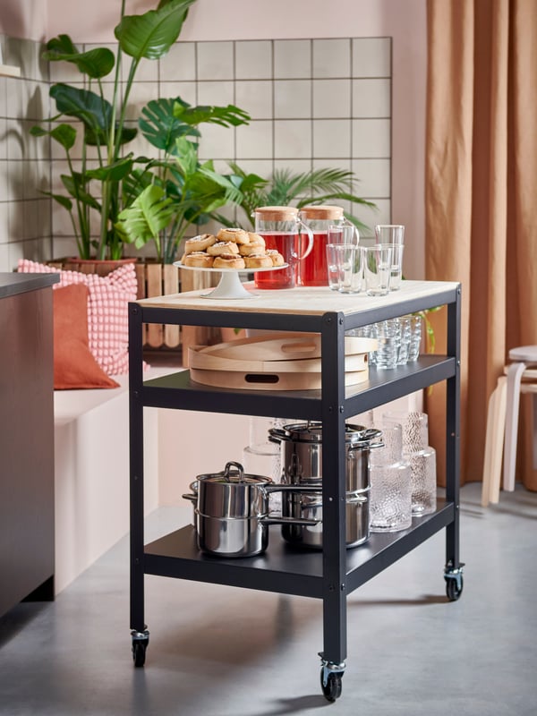 A black kitchen trolley with wooden top is standing by a bench with cushions in different colours on. Behind the bench is a tall green plant.