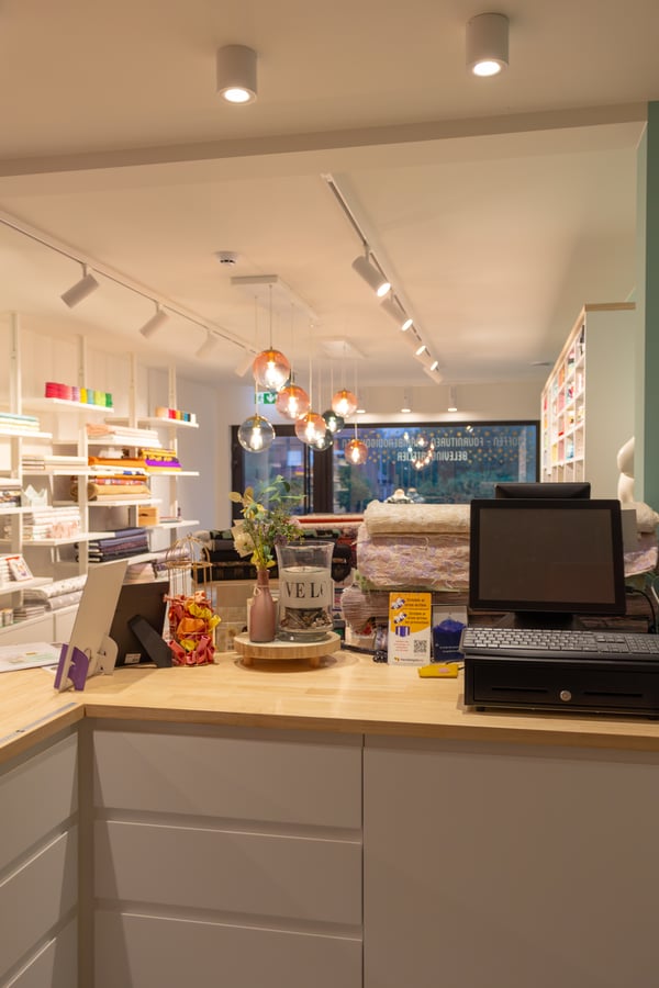 Een moderne winkelbalie in een lichte stoffenwinkel, met een kassa, laptop, kleine producten en zicht op rekken vol kleurrijke stoffen en hanglampen.