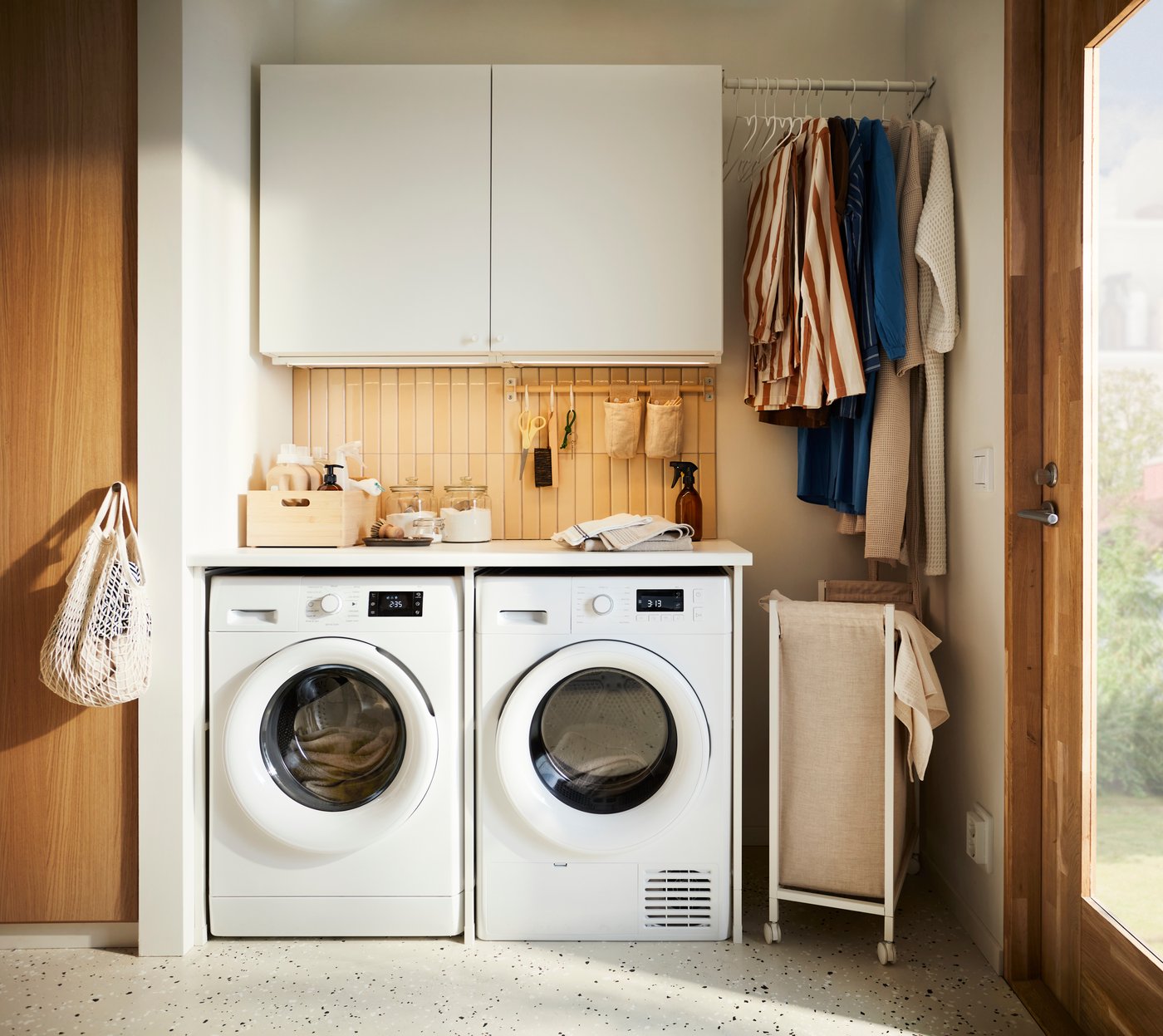 Een compacte ENHET wasruimte toont twee wasmachines onder een werkblad met houten wandpaneel, opbergkasten en kleding die langs de zijkant hangt.