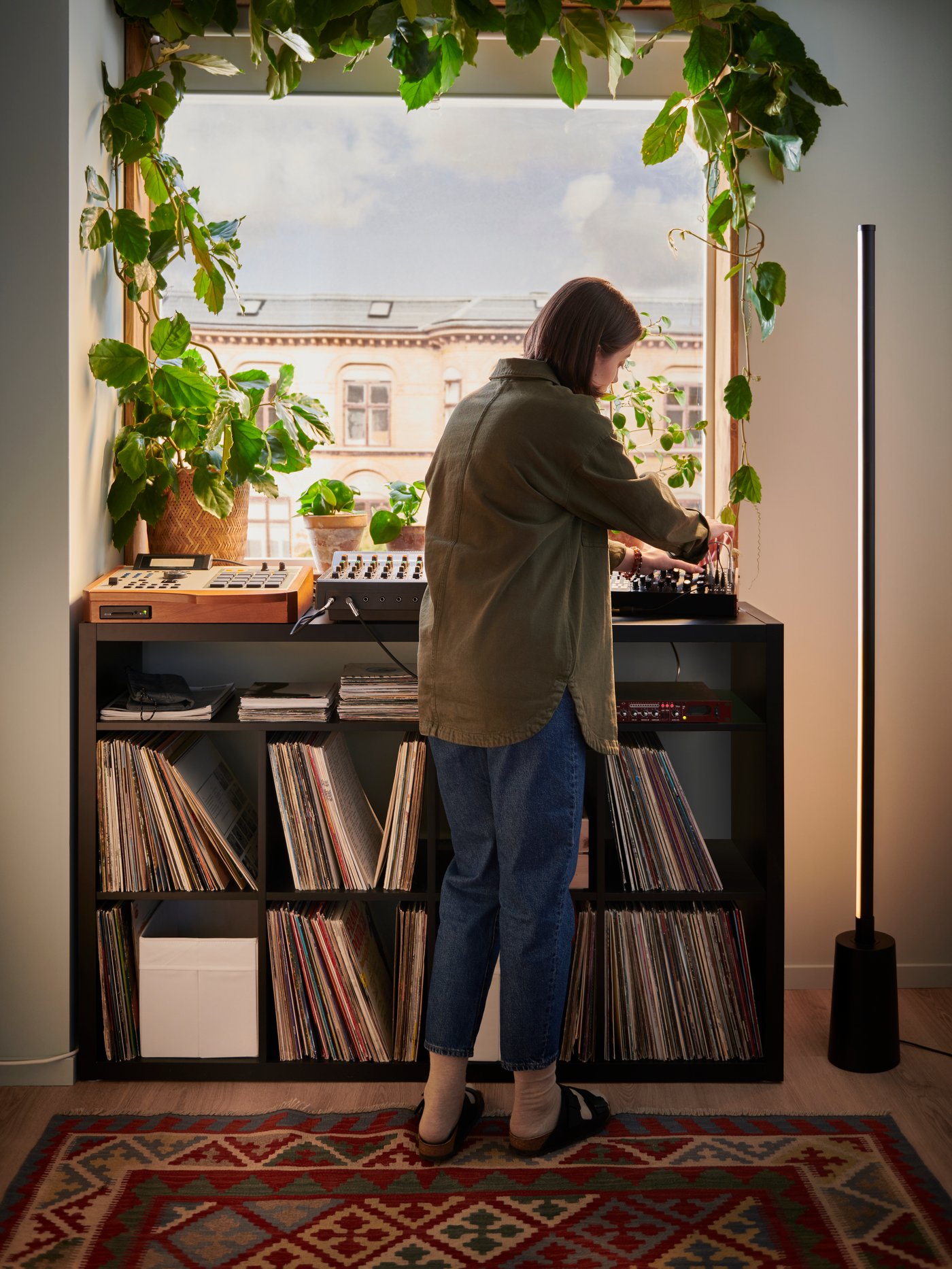 A woman standing at the black OBEGRÄNSAD shelving unit mixing music, standing on a rug with a plant-framed window behind her.