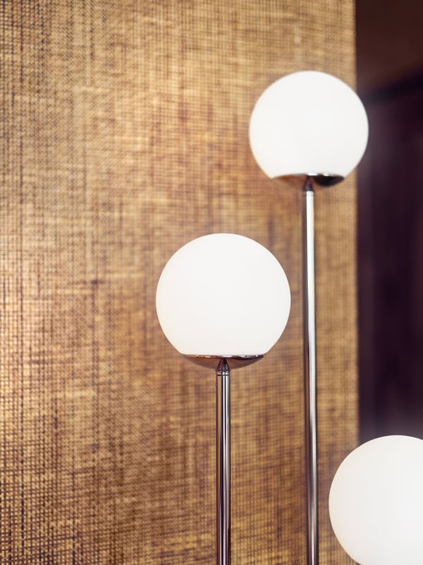 A chrome-plated SIMRISHAMN floor lamp with three round opal white shades placed in front of a light brown wall.