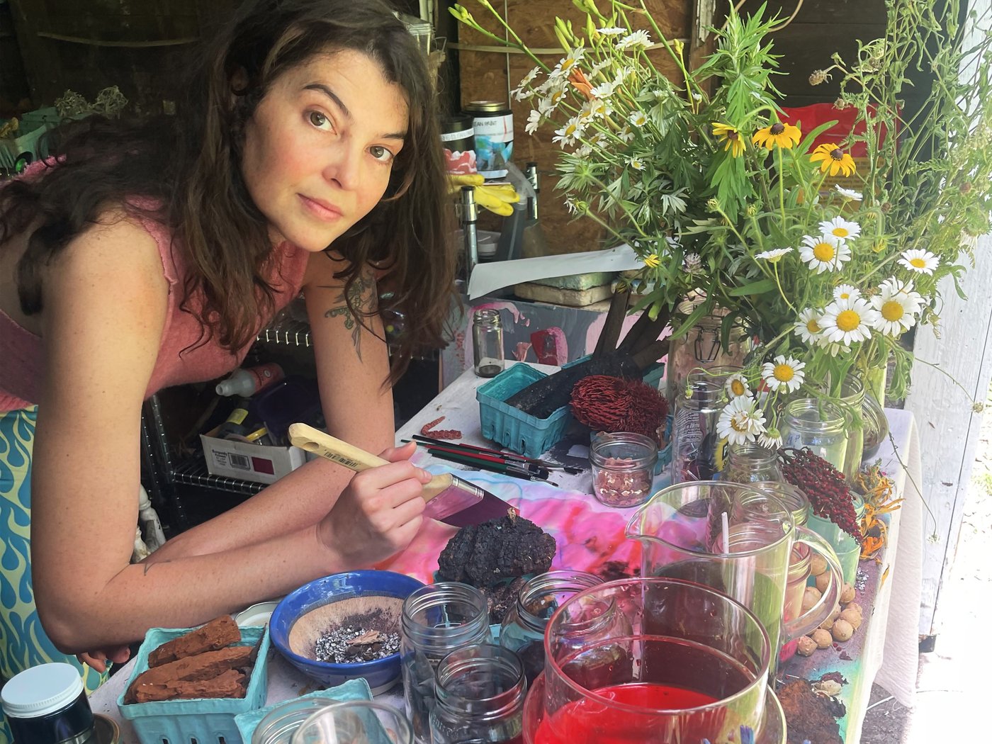 A woman is leaning over a desk full of glass jars and flowers. She is using a paintbrush to dye a piece of textile the colour pink.