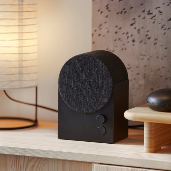 A black NATTBAD Bluetooth speaker with a rounded shape and two buttons on the front is standing on top of a pine cabinet.