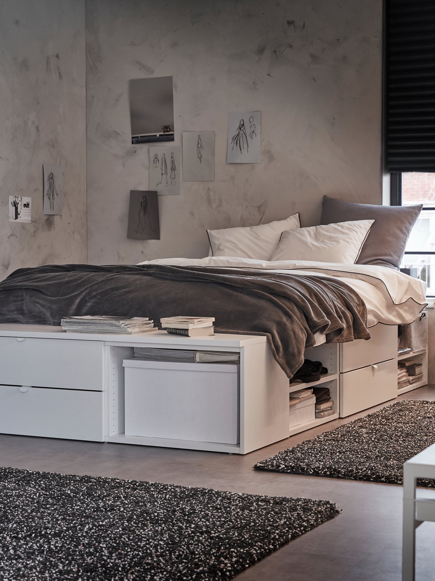 A bedroom with a white PLATSA bed with drawers, shelves and a bench stands by a window with a dark grey roller blind.