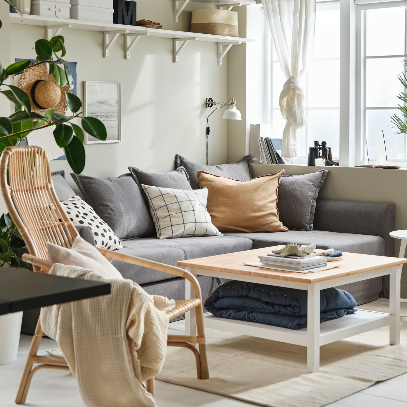 Dans un séjour cosy, canapé HOLMSUND gris à côté d’une fenêtre sous des étagères blanches, et fauteuil en rotin.