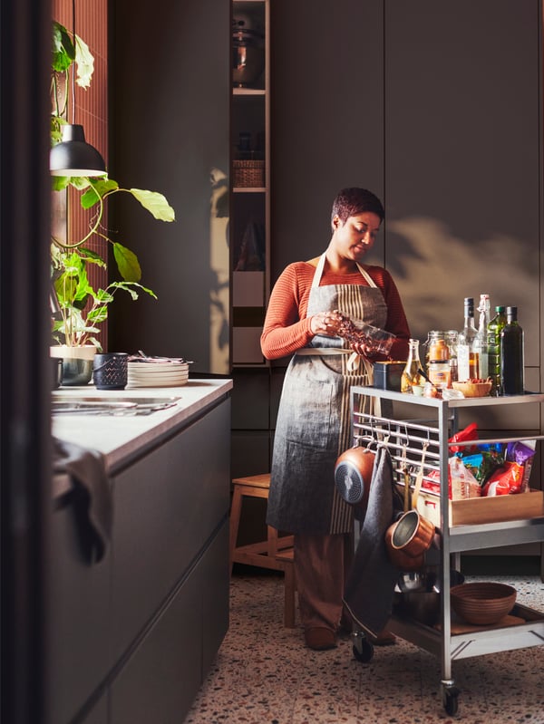 Une femme est debout dans une cuisine à côté d’une desserte KUNGSFORS. Elle a un ingrédient à la main et une porte VOXTORP est ouverte derrière elle.