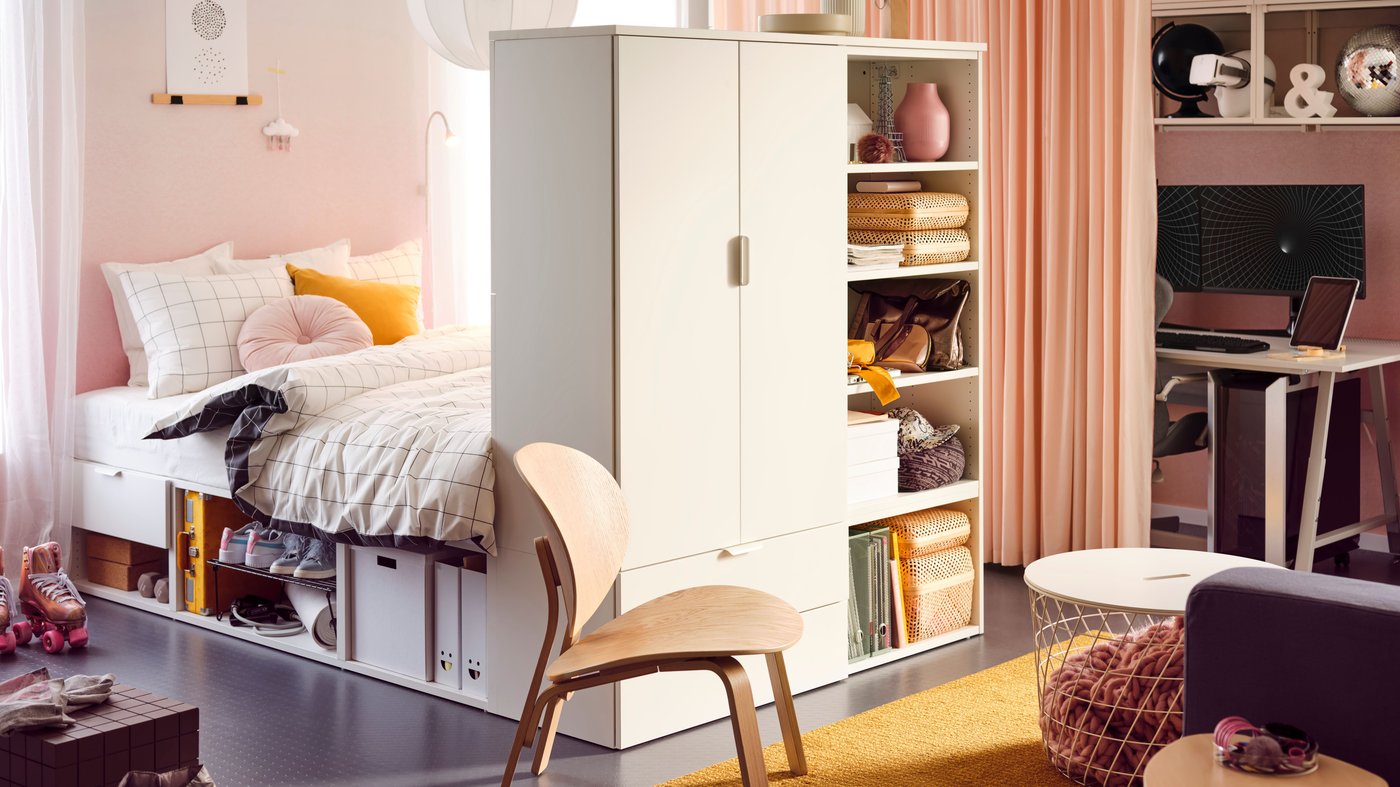 In der Mitte des Zimmer stehendes, weisses PLATSA Bett mit Schrankaufbau am Fussende zur Raumtrennung. Blass pinke MOALISA Vorhänge an der Wand die einen Arbeitsplatz verbergen.