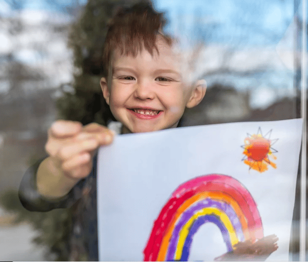Enfant présente un dessin d’un arc-en-ciel et d’un soleil derrière une fenêtre.