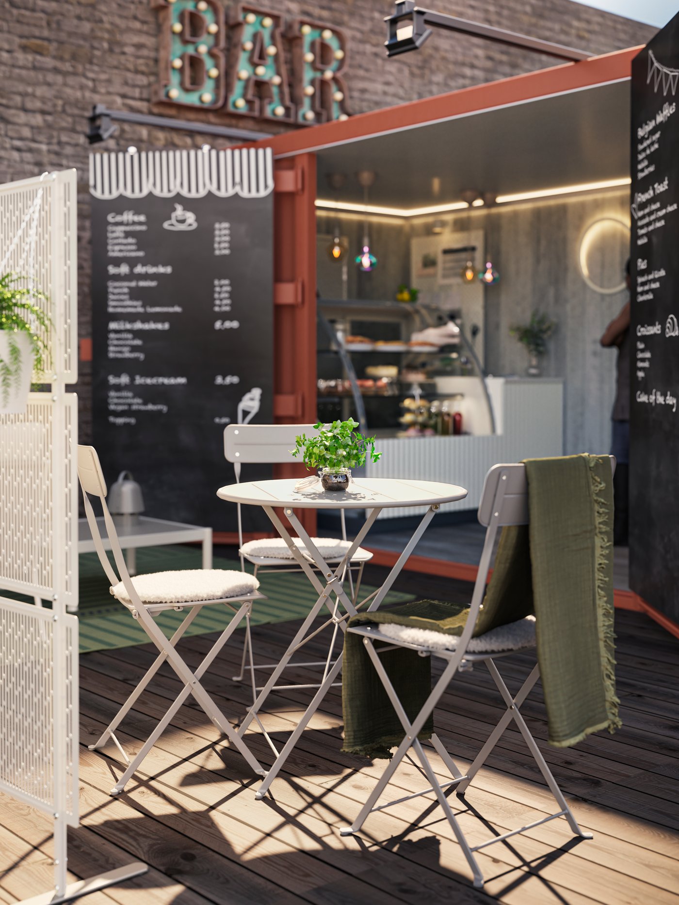 Kleiner Außenbereich einer Container-Bar mit rundem, weißem Klapptisch und zwei weißen KLAPPSTÜHLEN mit Sitzkissen auf Holzdeck, einem grünen Tuch über der Stuhllehne und einer Pflanze als Deko, vor einer offenen Theke mit Menütafeln im Hintergrund.