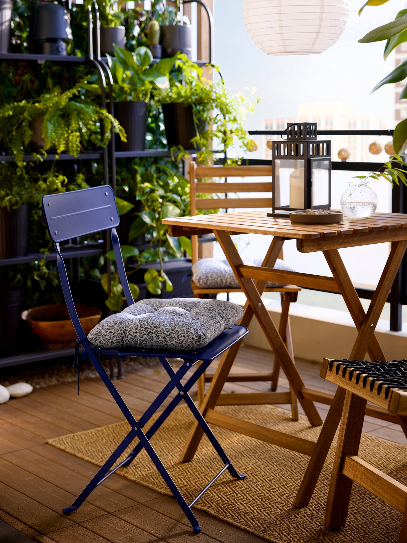 A blue KLÖSAN outdoor chair cushion with a floral pattern is tied onto a blue metal folding chair in a plant-filled balcony.