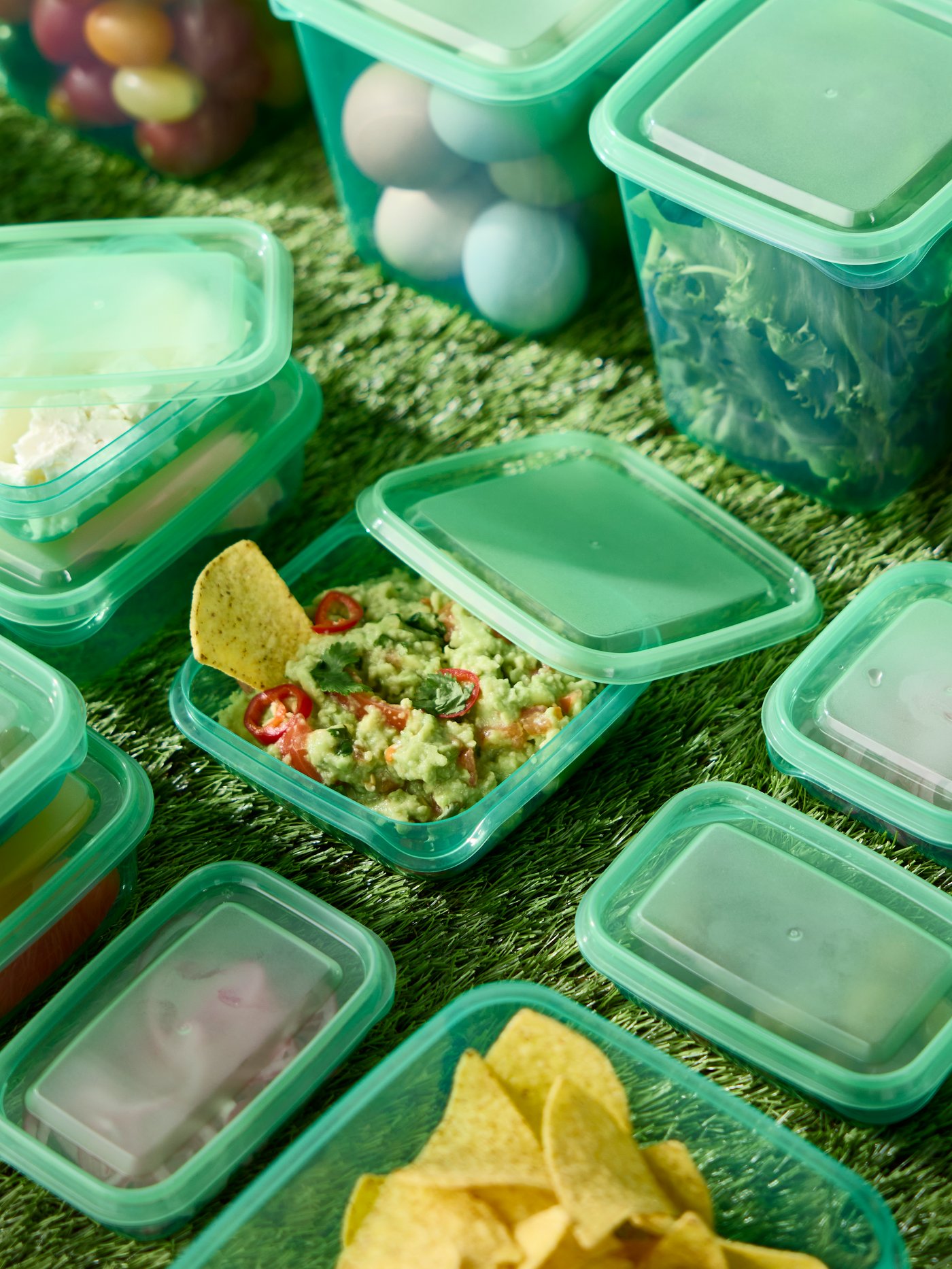 Green PRUTA food containers with lids are placed on a lawn and they are filled with tortilla chips, eggs and vegetables.