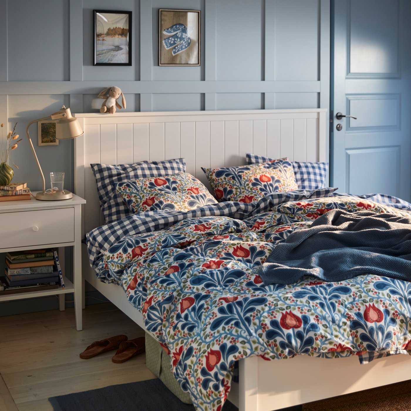 Une chambre aux murs peints en bleu, avec une parure de couette à motifs floraux SÄTERVIDE sur un lit blanc, à côté d'une table de chevet blanche surmontée d'une lampe.