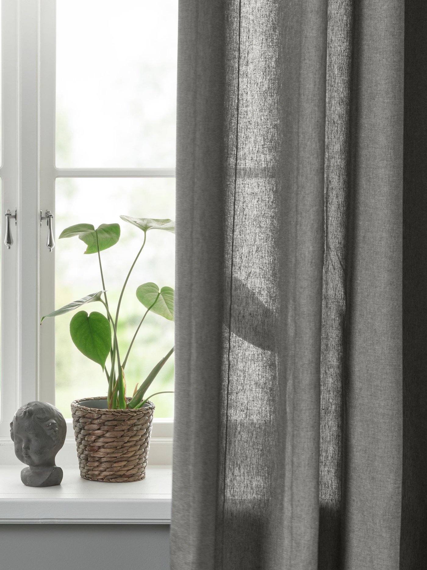 1 par de cortinas LENDA gris oscuro con tirantes y en el costado de la cortina una planta apoyada en el canto de la ventana.