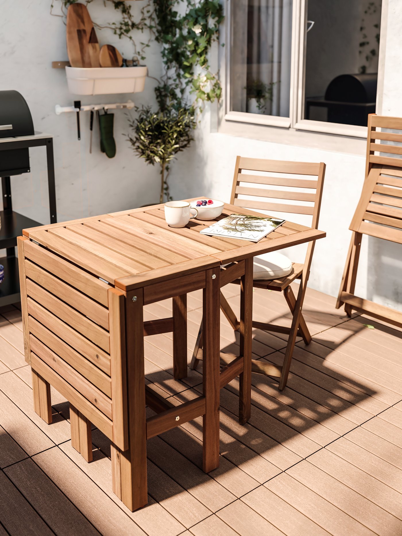 Una mesa de madera ÄPPLARÖ con un extremo plegado, frente a una silla plegable ÄPPLARÖ, en una terraza con piso de madera y muros blancos. Frente a la silla y sobre la mesa hay una revista, un tazón GLADELIG y un bowl de desayuno.