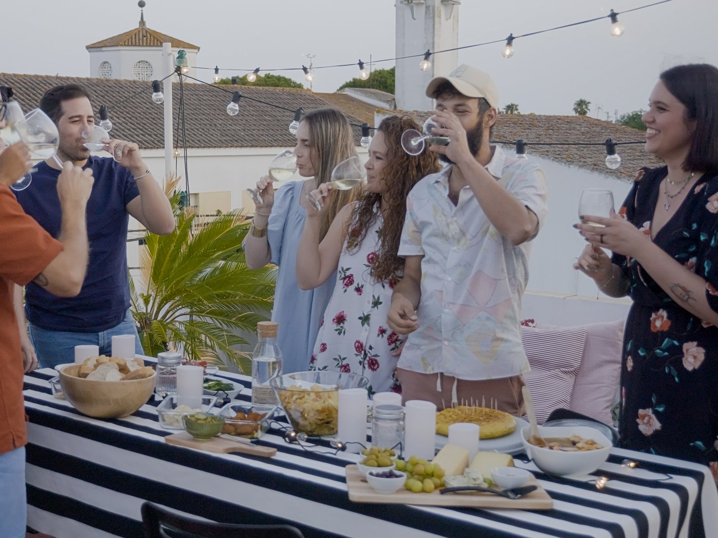 A group of young friends are cheerfully eating, drinking and talking, while standing at a buffet table full of food on an outdoor terrace.