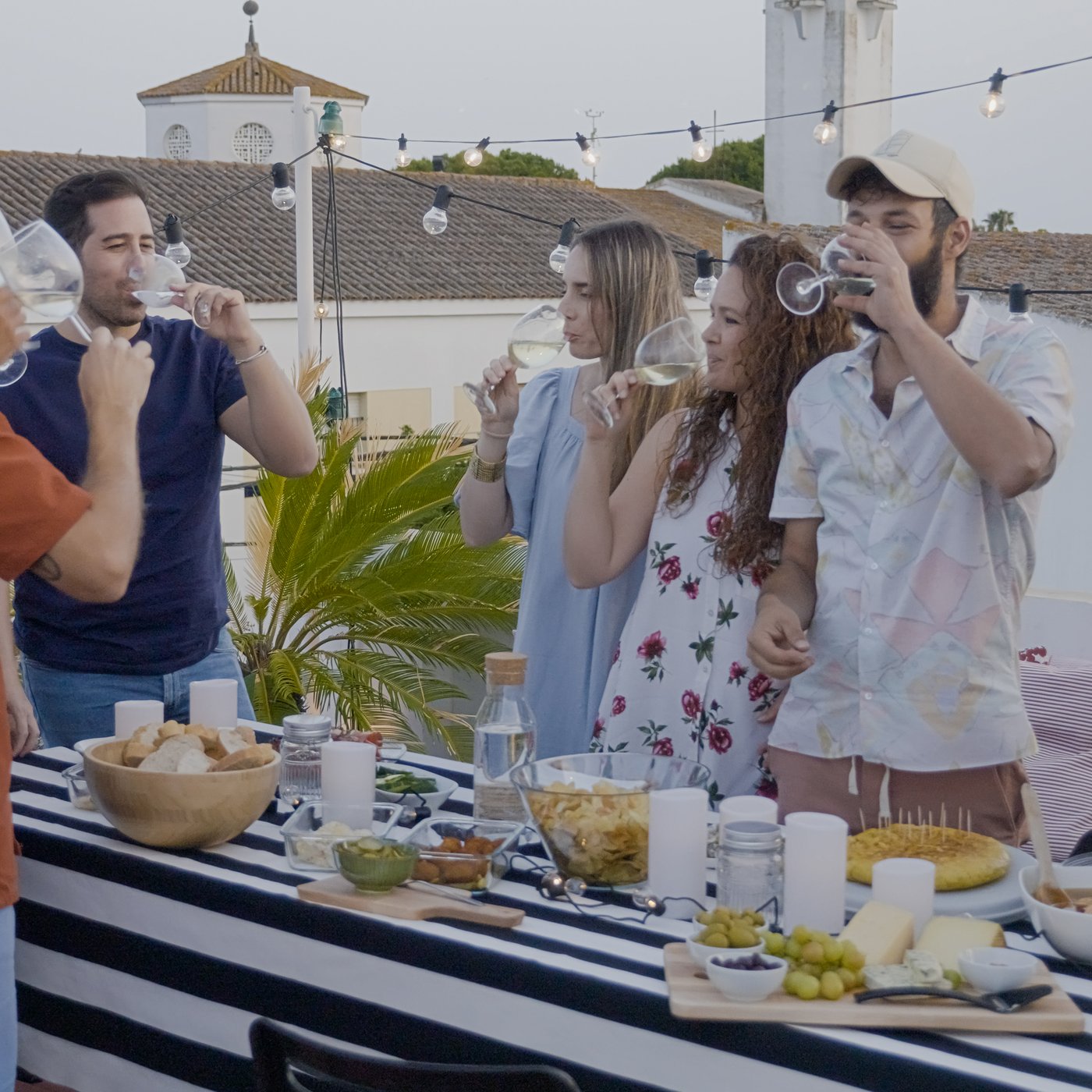 A group of young friends are cheerfully eating, drinking and talking, while standing at a buffet table full of food on an outdoor terrace.