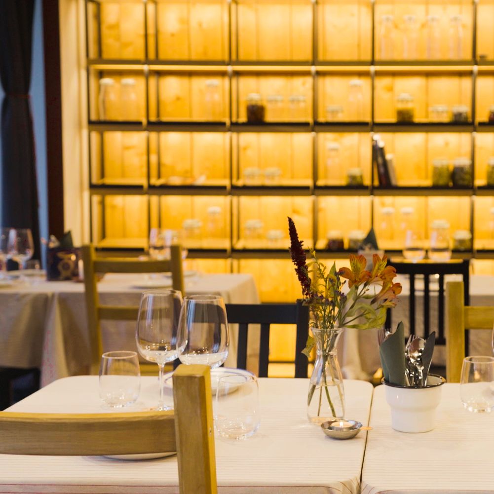 Gedekte tafels met bloemen, glazen en potjes in sfeervol restaurant. Achterwand met houten rekken en potten in warme, gezellige verlichting.