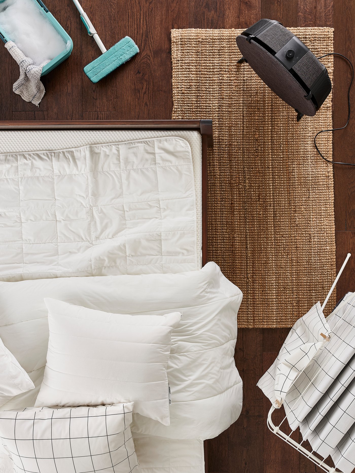 Una cama con ropa de cama blanca deshecha, ropa de cama sobre un tendedero y un purificador de aire STARKVIND redondo negro sobre una alfombra natural.